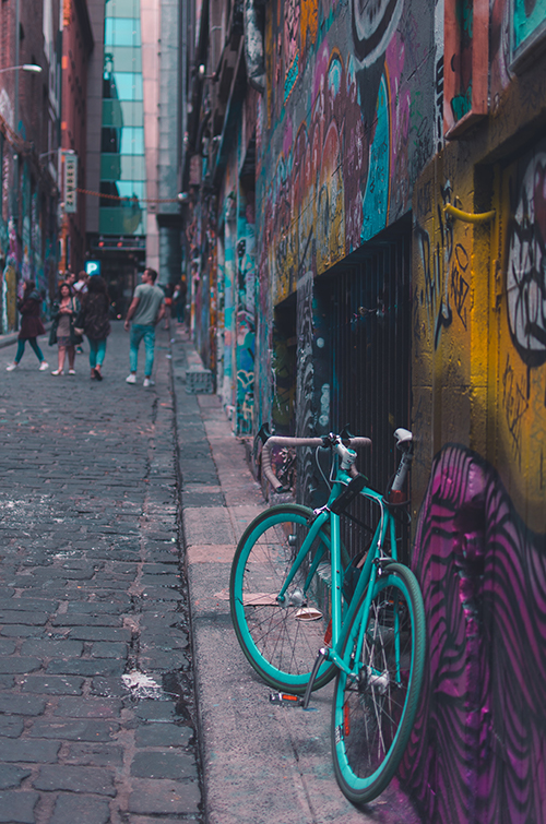 Bicycle leaning on a wall in a narrow alley with mural painting. Four people are walking in the distance.